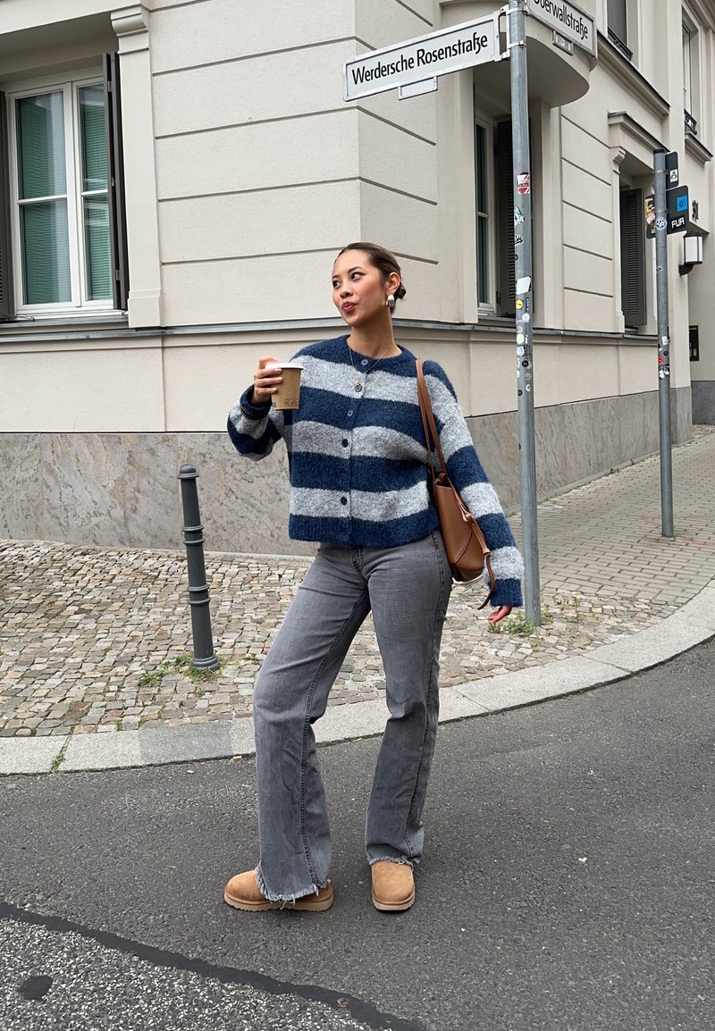 Gehaakte blauwe en grijze gestreepte cardigan met knopen, grijze jeans en tan enkelboots. Houdt een koffiekop vast, staande op een straathoek.