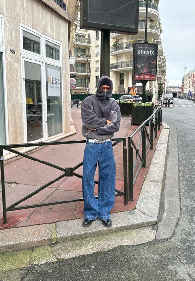 Sudadera de felpa gris y sobredimensionada con cubierta para la cara, combinada con unos vaqueros anchos de mezclilla azul y zapatos negros. Entorno urbano al aire libre con carteles.