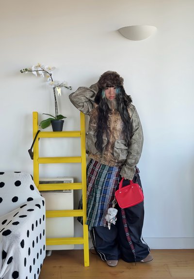 Chaqueta de camuflaje con bolsillos, falda de cuadros en varias colores, pantalones negros grandes, sombrero de piel marrón, bolso rojo, escalera amarilla al fondo.