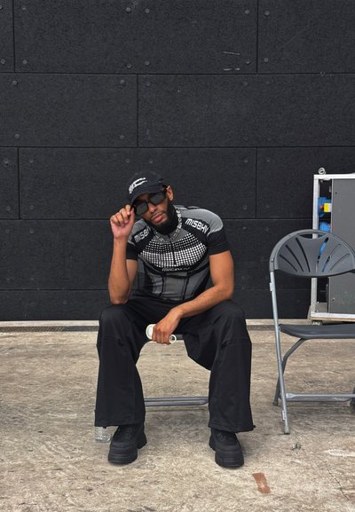 Gorra negra, gafas de sol espejadas, camisa ajustada con estampado y pantalones negros anchos. Sentado junto a una silla de metal sobre una superficie gris.