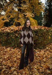 Jeune femme portant un pull marron à motifs losanges et un pantalon en cuir, debout sur des feuilles d'automne près d'une haie et d'arbres d'érable jaunes.