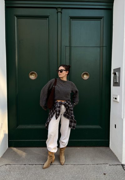 Mujer con gafas de sol, suéter gris corto, camisa de cuadros anudada, pantalones blancos y botas color canela, de pie frente a una gran puerta doble verde.