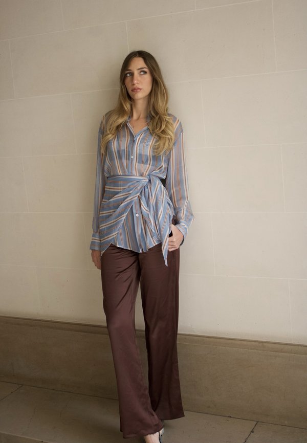Femme debout contre un mur beige portant un chemisier rayé bleu et orange avec taille nouée et pantalon marron à jambes larges.