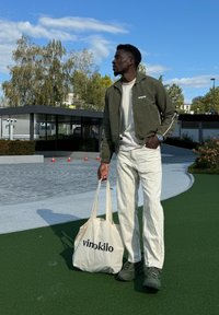 Πράσινο bomber jacket με λευκές λεπτομέρειες, κρέμα πουκάμισο, ανοιχτόχρωμο παντελόνι και γκρίζα αθλητικά παπούτσια. Κρατάει μια μπεζ τσάντα με εκτύπωση "vinokilo".