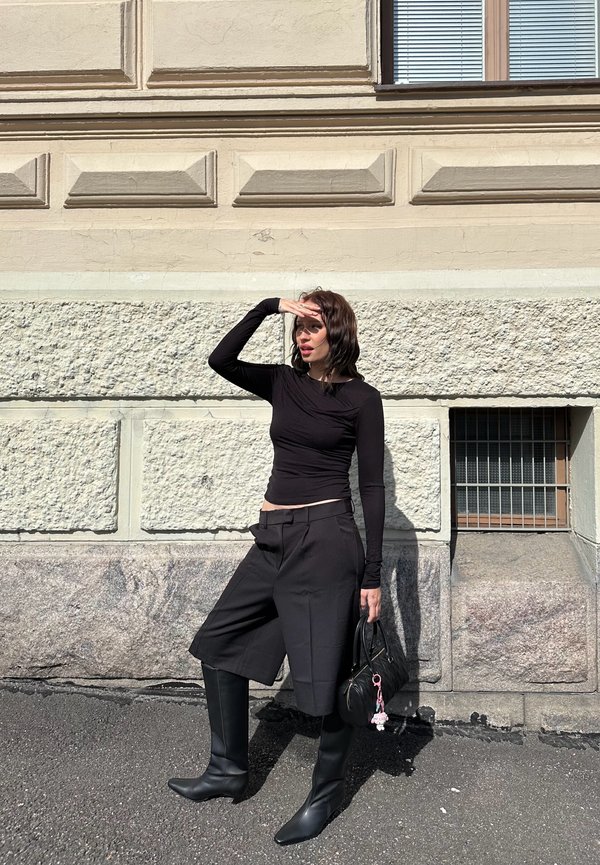 Črn dolgi rokav top, hlače visoke pasu in črne čevlje do kolen s koničastim vrhom. Drži majhno črno torbico z obeskom. Urbano okolje z zidom iz kamna.