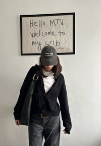 Personne portant une casquette noire et un cardigan se tient sous un panneau encadré où il est écrit « Hello MTV welcome to my crib » sur un mur blanc uni.