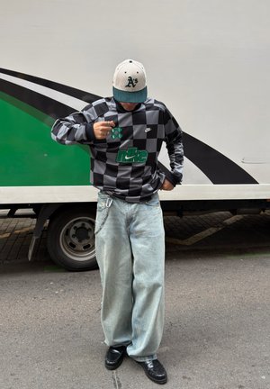 Persona con una sudadera de Nike a cuadros en negro y gris, vaqueros anchos de color azul claro, zapatos negros y una gorra de béisbol blanca y verde, de pie al aire libre.