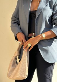 Blazer cinza sobre uma blusa preta, segurando uma bolsa tecida em tom bege suave. Anéis dourados notáveis e um relógio com mostrador retangular. Calças escuras.