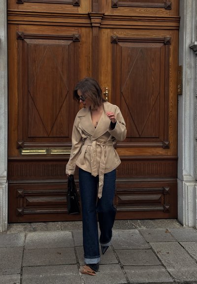 Mujer con chaqueta beige con cinturón, vaqueros oscuros, botas negras y gafas de sol caminando sobre un pavimento de piedra frente a grandes puertas dobles de madera.