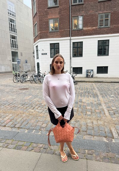 Joven mujer de pie en una calle de adoquines sosteniendo un bolso tejido naranja, vistiendo una blusa blanca de manga larga, una falda negra y chanclas.