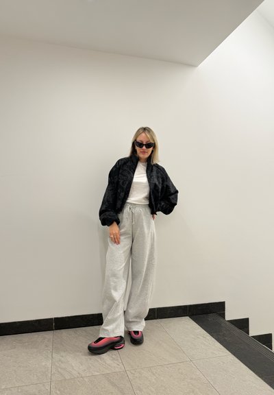 Chaqueta bomber negra con textura estampada, camiseta blanca, pantalones de chándal gris claro y zapatillas chunky negras y rosas. De pie en un espacio minimalista.