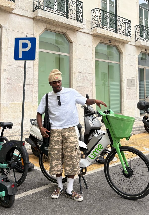 Muž s béžovou čiapkou a maskáčovými šortkami stojí vedľa zaparkovaného elektrického bicykla Lime na mestskej ulici blízko parkovacieho značenia.