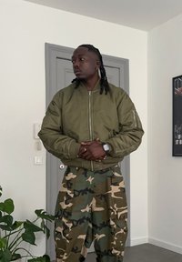 Veste bomber vert olive avec une poche zippée, associée à un pantalon ample à motif camouflage. Cheveux foncés tressés et une montre visible.