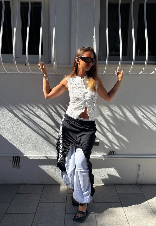 Frau mit Sonnenbrille, ärmellosem weißen Top, geschichteter schwarzer und grauer Hose, posiert mit Händen, die vertikale Metallstangen an einer sonnenbeschienenen Wand halten.