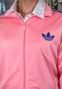Rožinė satino sportinė striukė su užtrauktuku priekyje, balta apykakle ir mėlynu adidas logotipu kairėje krūtinės pusėje; ant pečių yra mėlynos juostos.