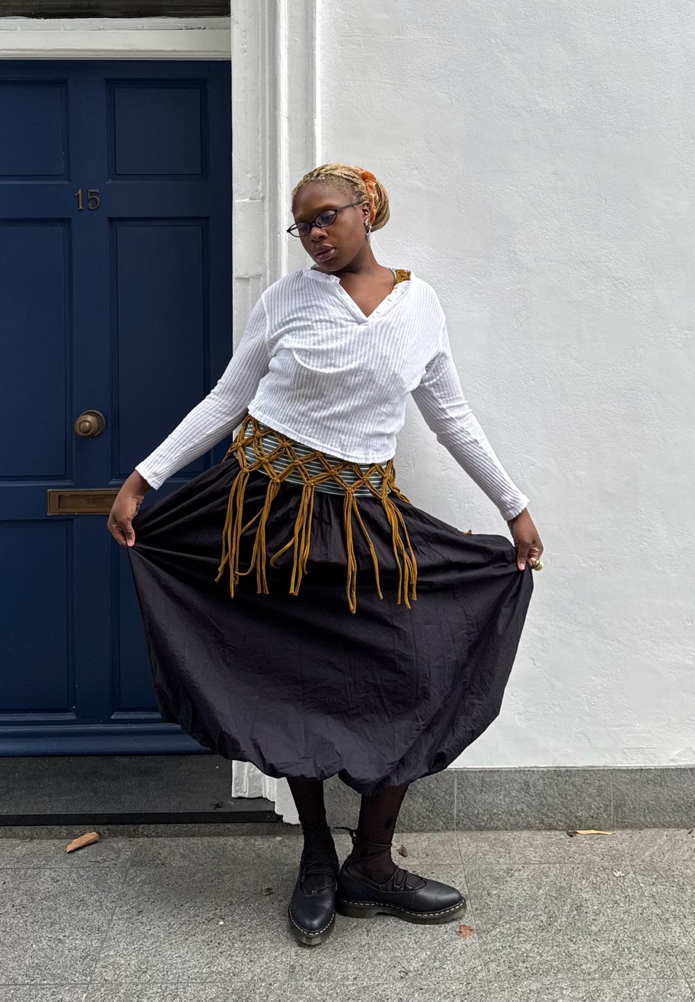 Schwarzer langer Rock mit Fransen, weißes, geripptes Langarmoberteil und schwarze Schuhe. Stehend vor einer weißen Wand und einer blauen Tür.