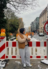 Άτομο που φοράει ένα καπιτονάτο καφέ γιλέκο και γκρίζες φόρμες στέκεται δίπλα σε μια κατασκευαστική περίφραξη με πορτοκαλί προειδοποιητικά φώτα και αστικό φόντο.