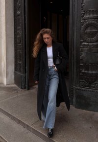 Svart lang frakk, hvit t-skjorte og lyseblå vide jeans. Svart håndveske, stiletto-støvler. Betongtrinn og dekorativ dør i bakgrunnen.