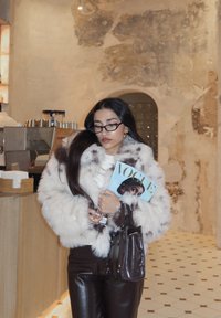 Γυναίκα με λευκό και καφέ γούνινο παλτό κρατά περιοδικό Vogue και καφέ δερμάτινη τσάντα σε ένα ρουστίκ καφέ με ανάγλυφους τοίχους και πλακάκια στο δάπεδο.