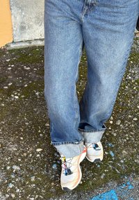 Ténis desportivos brancos e bege com detalhes multicoloridos e atacadores pretos; jeans de ganga azul acinzentado com bainha; superfície exterior de cascalho.