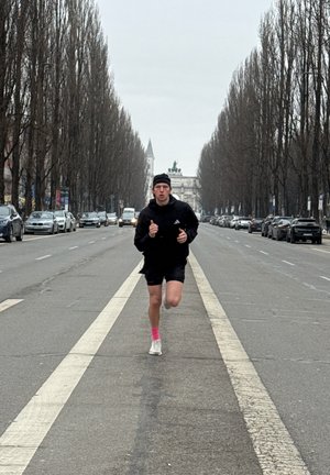 Joven con ropa deportiva negra corriendo en el centro de una amplia calle de la ciudad bordeada de árboles, con coches aparcados a ambos lados.