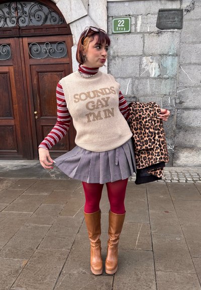 Persona con chaleco suéter que dice "SOUNDS GAY I'M IN", camiseta de manga larga a rayas rojas, falda plisada, medias rojas, sosteniendo un abrigo con estampado de leopardo junto a una pared de piedra.