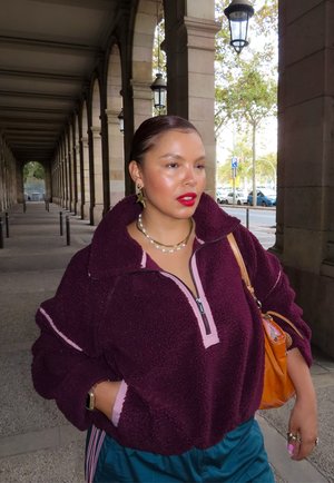 Sudadera de forro polar burdeos con cremallera y ribete rosa. Pantalones verdes, bolso naranja y pendientes en capas de perlas y estrellas.