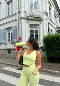 Lysegrønn crop top og leggings med tettsittende tekstur, kombinert med solbriller og svart veske, stående nær en bygning med blå persienner.