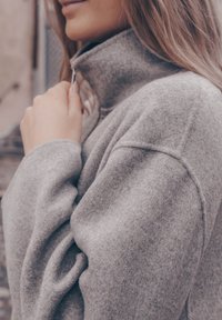 Γκρίζα φλις μπλούζα με φερμουάρ και ψηλό γιακά, με μαλακή υφή και ελαφρώς φουσκωτά μανίκια, που τονίζεται από μια λεία λεπτομέρεια φερμουάρ.