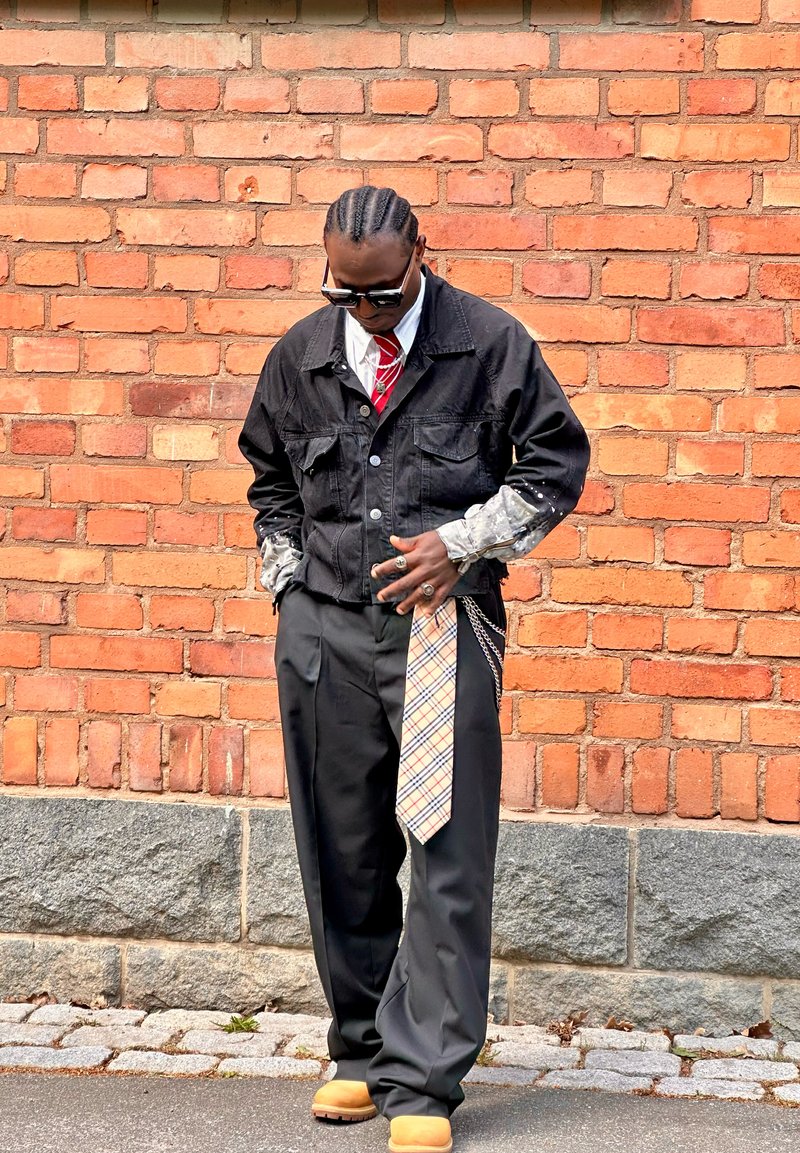 Homme avec des cheveux tressés portant des lunettes de soleil, une veste noire, une cravate à motifs suspendue au pantalon, une cravate rouge et des bottes beige, debout contre un mur en briques.