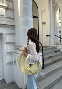 Bolsa amarela texturizada com padrão de crocodilo, apresentando um detalhe de fivela lateral. Usada ao ombro, combinada com uma blusa de manga longa branca.