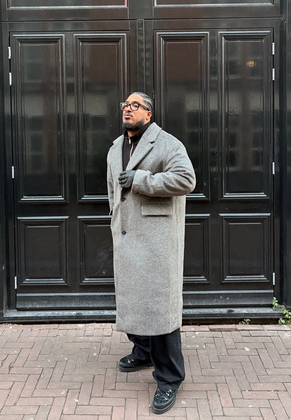 Muž s vrkočmi a okuliarmi stojaci pri vysokých čiernych dverách s panelmi, oblečený v dlhom sivom kabáte, čiernych rukaviciach a čiernych nohaviciach.