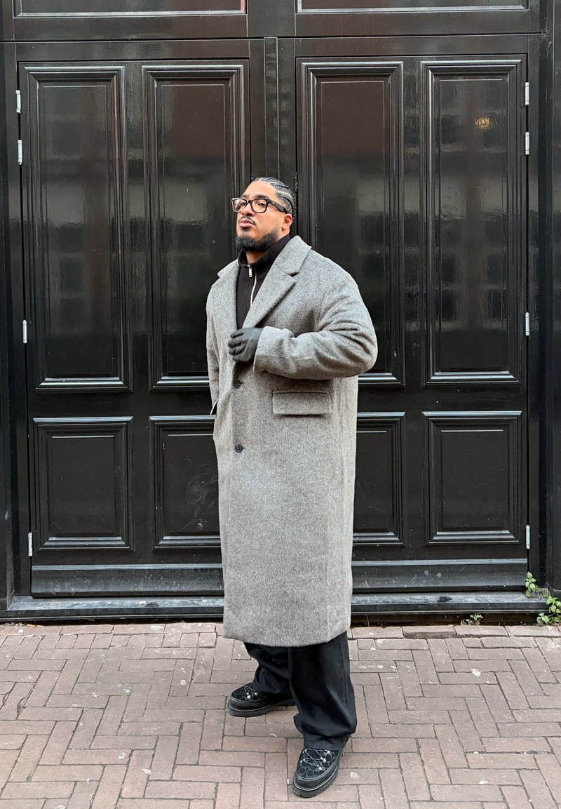 Homme aux cheveux tressés et lunettes, portant un long manteau gris, des gants noirs et un pantalon noir, debout contre une grande porte noire à panneaux.