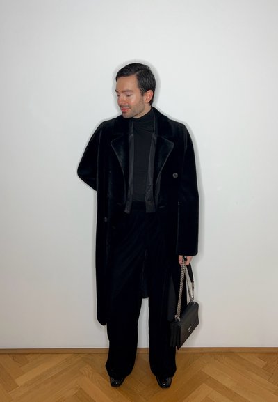 Abrigo de terciopelo negro con grandes botones, combinado con un cuello de tortuga negro y pantalones de pierna ancha. Lleva un bolso negro con una correa de cadena plateada.