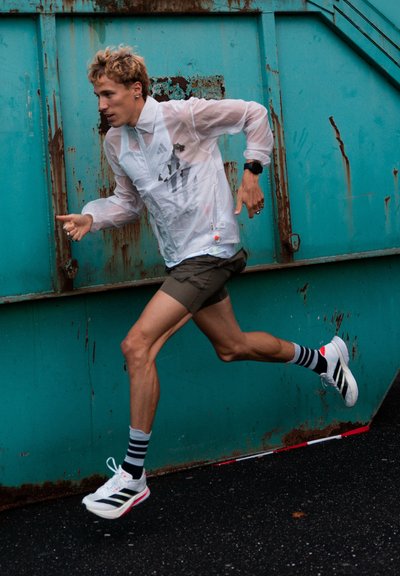 Figura masculina atlética con chaqueta blanca translúcida, pantalones cortos verdes y calcetas a rayas, corriendo contra un fondo texturizado color verde azulado.