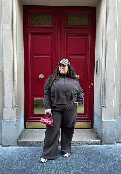 Sudadera cremallerada de forro marrón y pantalones a juego, bolso de mano con estampado de cocodrilo rojo y gorra de estampado de leopardo. De pie cerca de puertas dobles rojas.