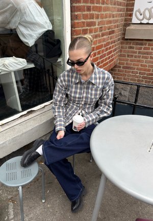 Joven mujer con gafas de sol y una camisa de cuadros sentada afuera en una mesa redonda sosteniendo una taza de café con una pierna apoyada en un taburete.