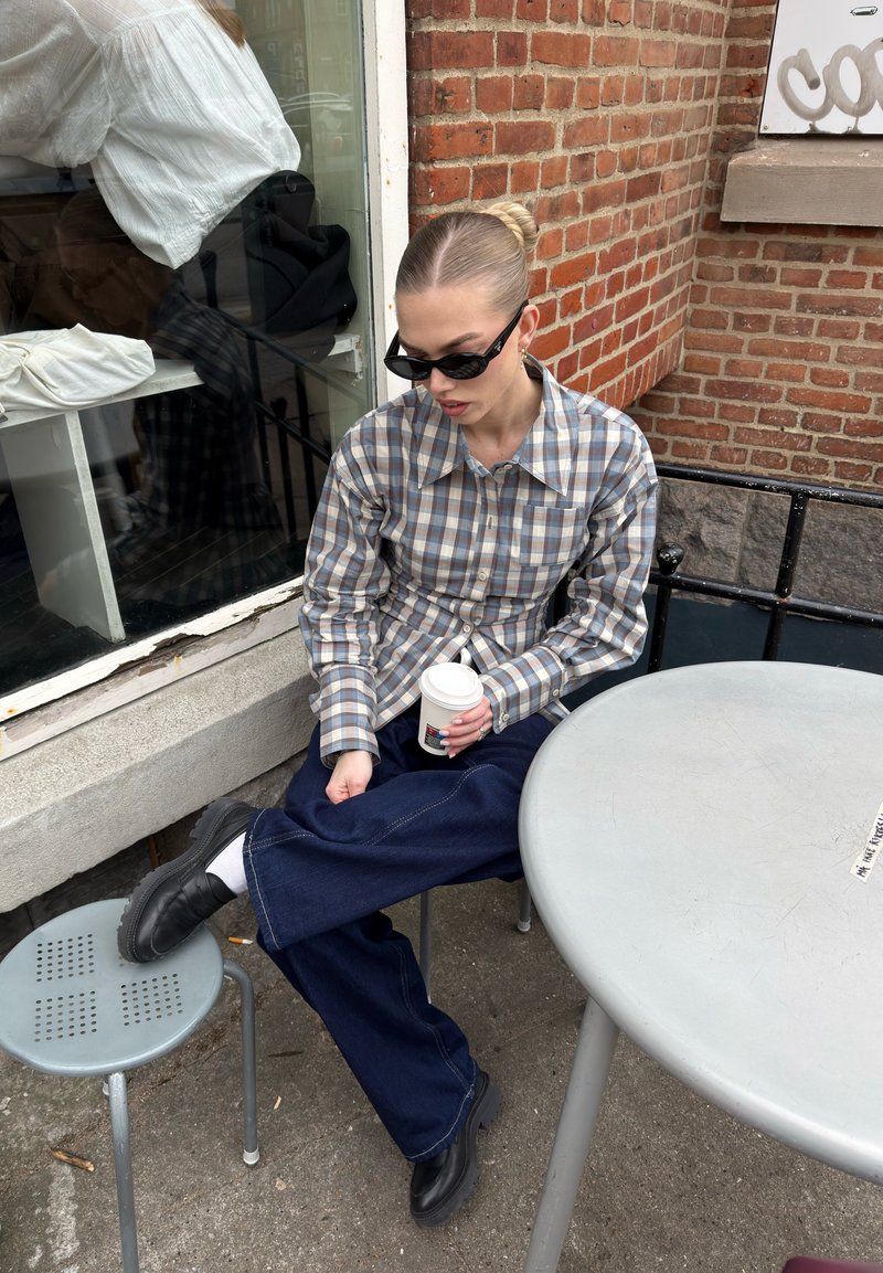Jeune femme portant des lunettes de soleil et une chemise à carreaux assise dehors à une table ronde, tenant une tasse de café avec une jambe reposant sur un tabouret.