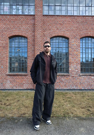 Hombre con gafas de sol, chaqueta negra y pantalones anchos, de pie sobre el césped frente a un edificio de ladrillo rojo con grandes ventanas arqueadas.