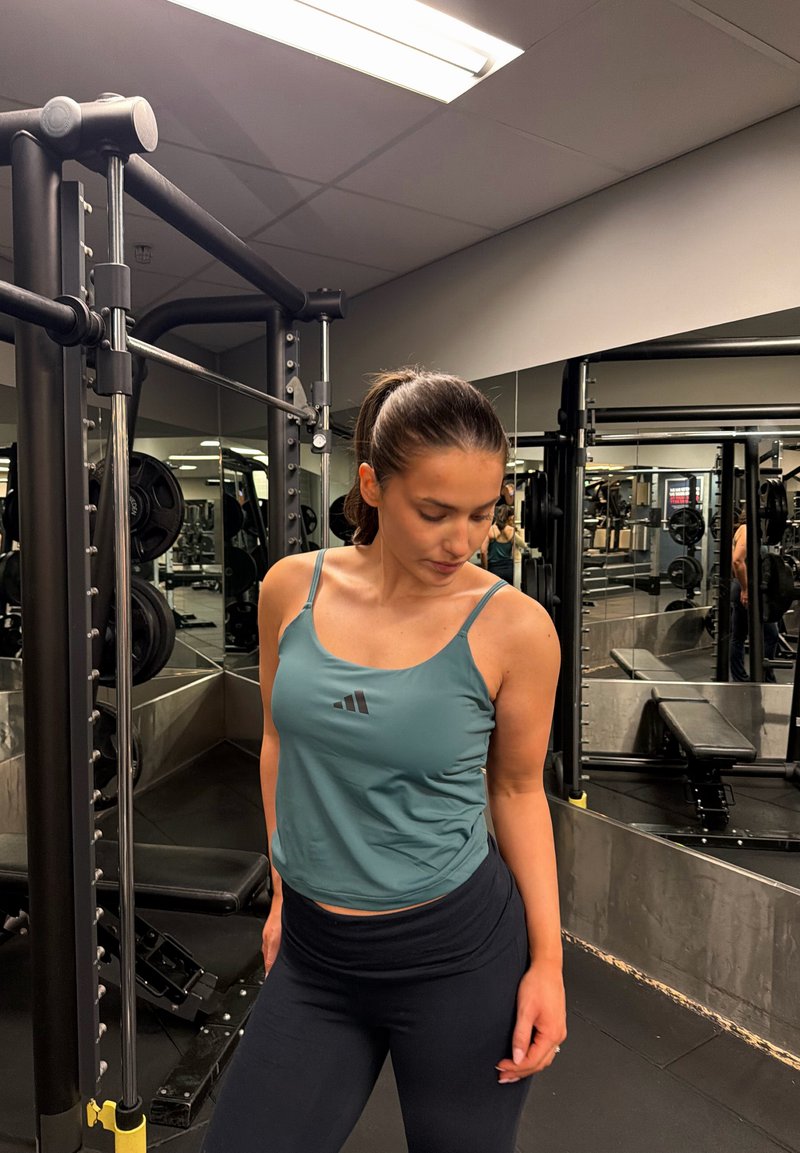 Femme aux cheveux bruns attachés en queue de cheval, portant un débardeur turquoise et des leggings noirs, debout à l'intérieur d'une salle de sport près d'équipements de musculation et de miroirs.