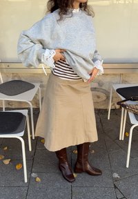 Siv širok pulover z dodatki iz čipke, pod njim črtasta majica z dolgimi rokavi, bež krilo na zvonček in temno rjavi škornji do kolen.