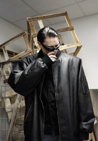 Blouson aviateur en cuir noir avec col montant, fermetures éclair latérales et poignets côtelés. Porté sur une chemise noire, sur un fond neutre.