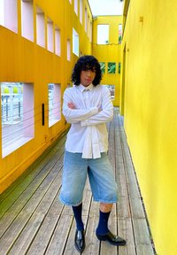 Witte button-up shirt met een kraag, gedeeltelijk ingestopt, gecombineerd met lichtblauwe denim shorts. Zwarte sokken en puntige zwarte schoenen. Heldergele achtergrond.