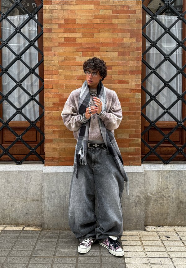 Personne aux cheveux bouclés et portant des lunettes, habillée d'un jean gris surdimensionné, d'un pull beige, d'une écharpe noire et blanche, et de baskets fleuries, debout contre un mur en briques.