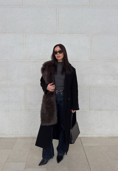 Mujer con gafas de sol que lleva un abrigo largo negro con una bufanda de piel gris, top gris oscuro, vaqueros de pierna ancha, botas negras de punta y un bolso negro.