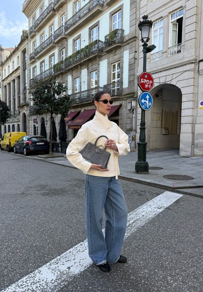 Mujer con gafas de sol que lleva una chaqueta cremosa y vaqueros holgados, sostiene un bolso gris, de pie en una calle de la ciudad con edificios históricos y señales de tráfico.