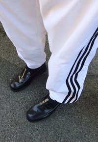 Persona, kas ģērbjas baltās biksēs ar melnām svītrām un melnu ādas slip-on apavu pāri, stāv uz asfalta virsmas.