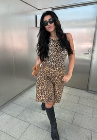 Top sem mangas em padrão de leopardo e calções, em tons de castanho e dourado. Botas pretas até ao joelho e óculos de sol. Joias douradas e um acessório suave em castanho.