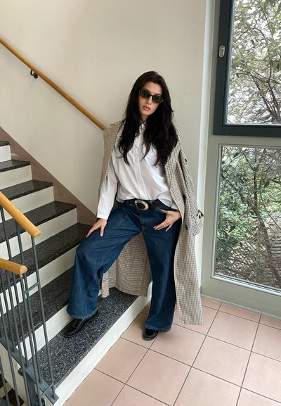 Modelo luciendo un abrigo trench de cuadros largo sobre una camisa blanca, combinado con jeans de pierna ancha azules y zapatos negros, posando en una escalera.