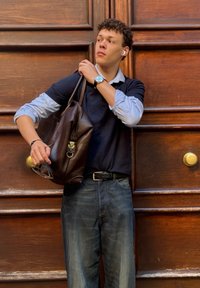 Zaino in pelle marrone con una chiusura argentata, indossato con un maglione navy e una camicia blu con colletto. Jeans scuri, orologio tondo e occhiali in mano.